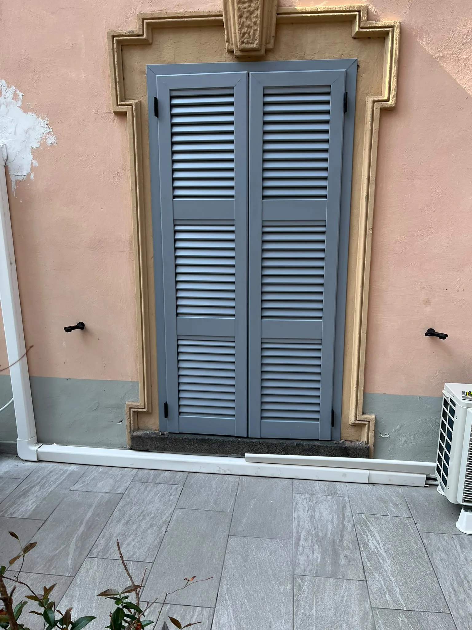 Persiane grigio-azzurre a due ante chiuse su un’apertura con cornice decorativa, sulla facciata rosa di un edificio a Milano.