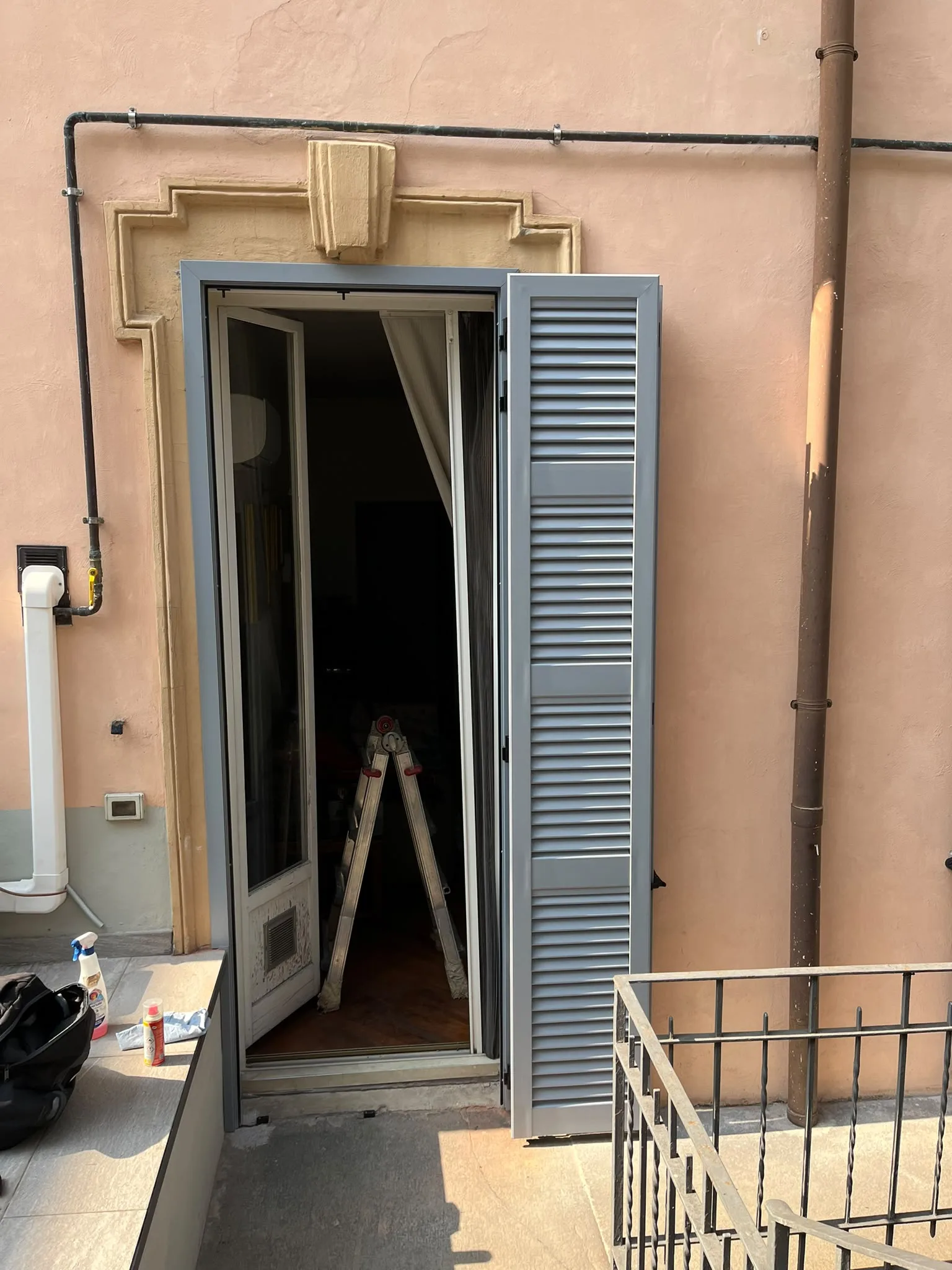 Una persiana grigio-azzurra aperta accanto a una porta-finestra sulla facciata rosa di un edificio a Milano.