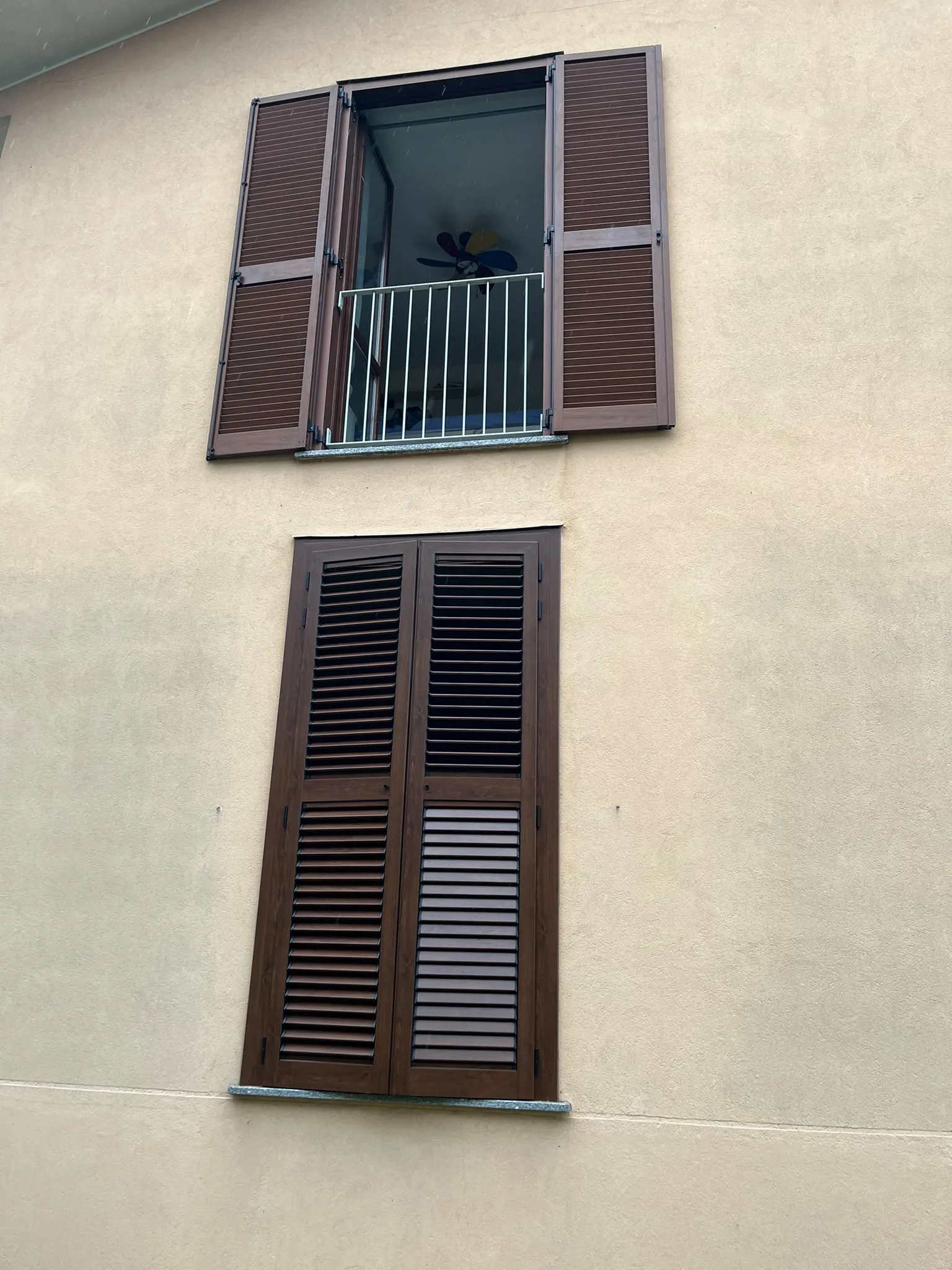 Persiane marroni a lamelle aperte sulla finestra superiore e chiuse su quella inferiore di una facciata chiara a Novara.