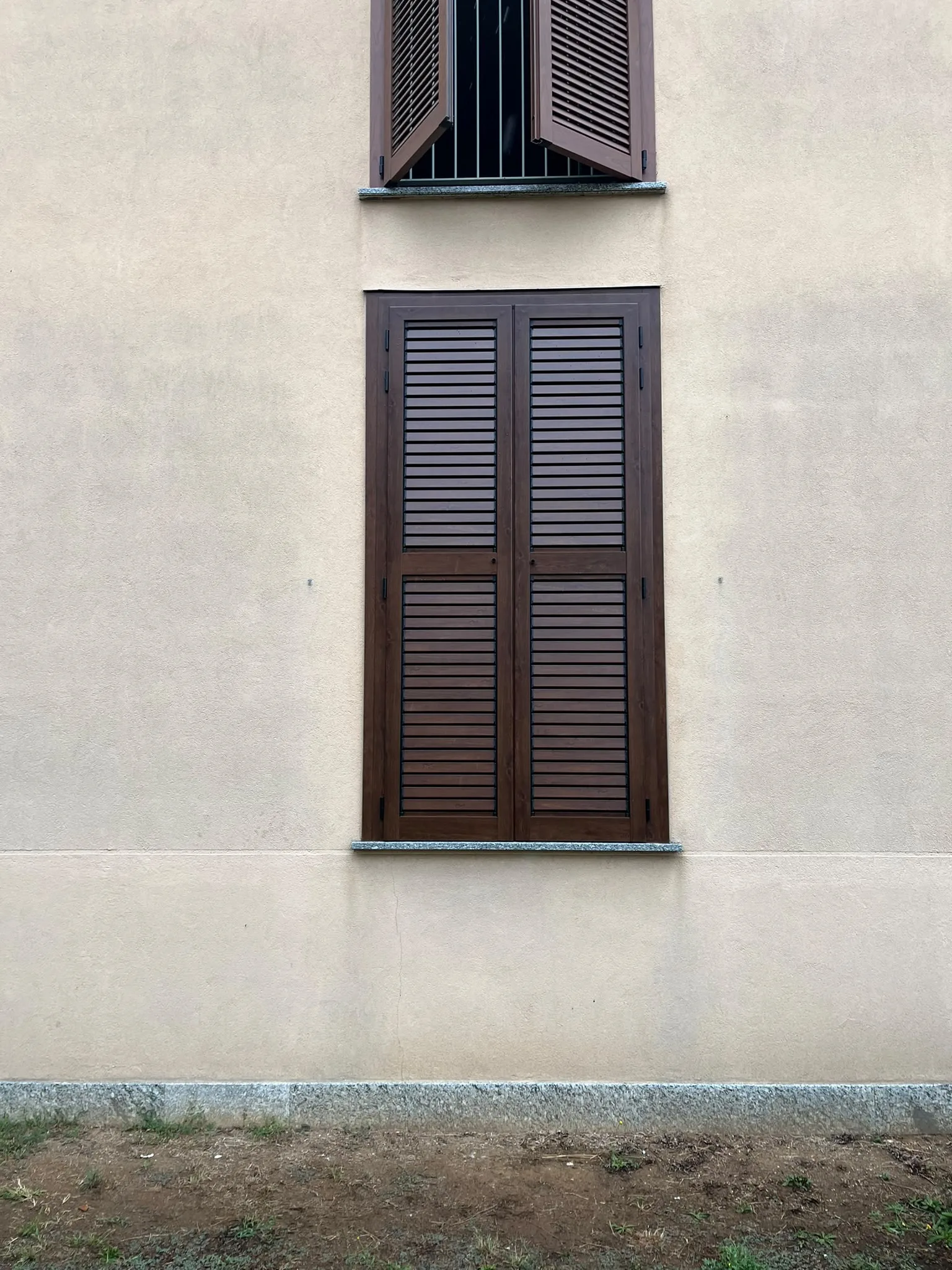Persiana marrone a due ante chiusa sulla finestra inferiore di una facciata chiara a Novara, con un’altra finestra in alto.