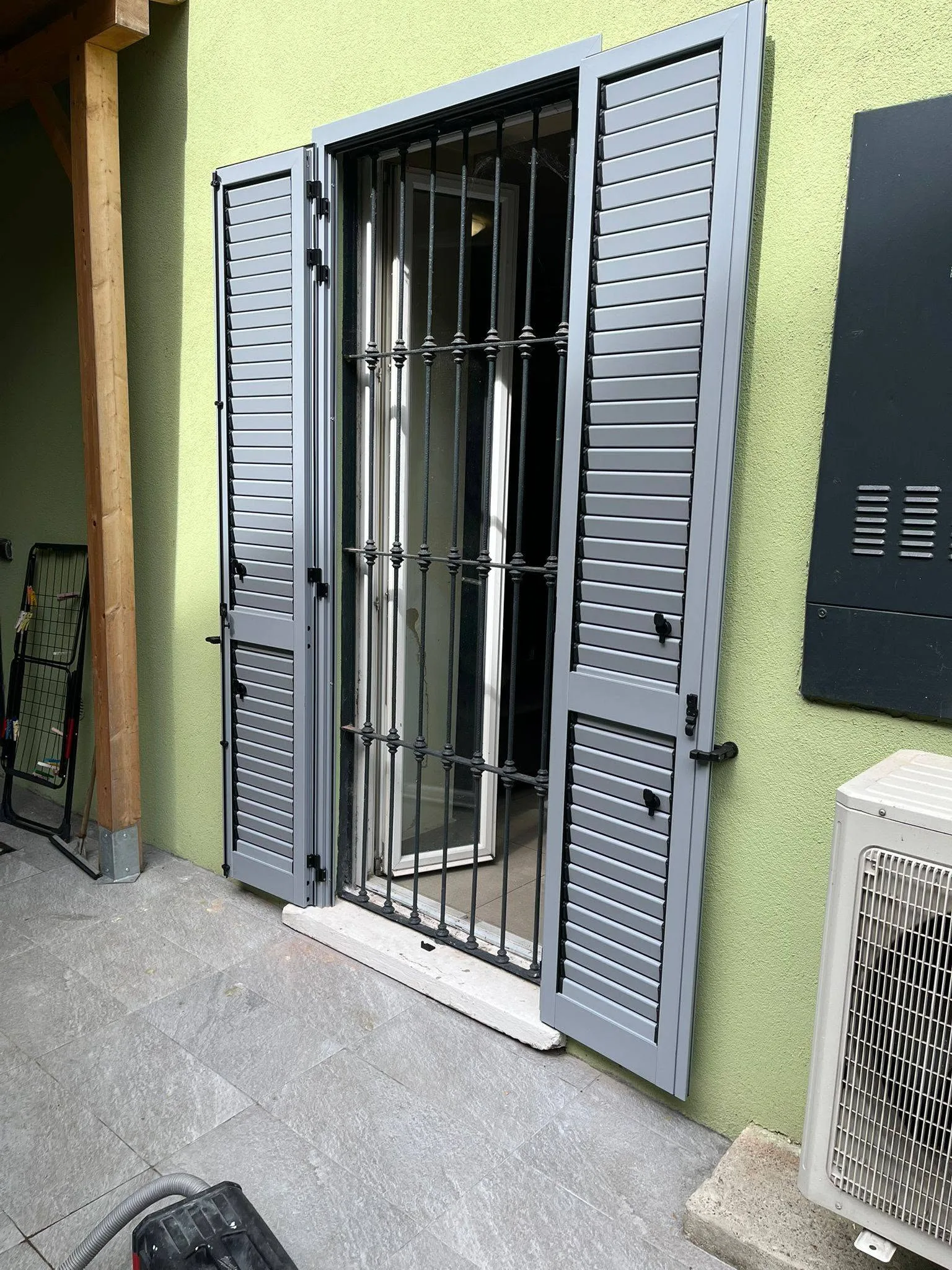 Due persiane grigie a lamelle aperte ai lati di una porta-finestra sulla facciata verde di una casa a Varese.